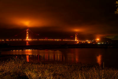Golden Gate Bridge illuminated at night with fog glowing orange and reflections on the bay