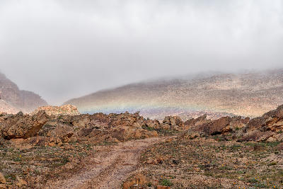 Remote gravel road winding through rocky sandstone terrain between Matjiesrivier and Wupperthal, with low cloud, mist, and a faint rainbow across the mountains.