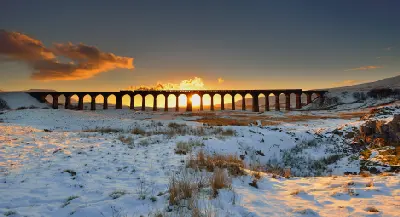 Steam locomotives crossing Ribblehead Viaduct at sunset, with golden sunlight shining through the stone arches above a snow-covered Batty Moss.