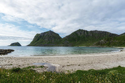View across Vik Beach on Vestvågøya in the Lofoten Islands, showing a sandy shoreline, calm Arctic water and steep green mountains rising from the coast in Norway.