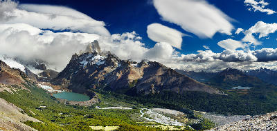 Wide panoramic landscape of Laguna Torre, river valley, and Mount Fitz Roy peaks under dramatic cloud formations in Patagonia, Argentina.