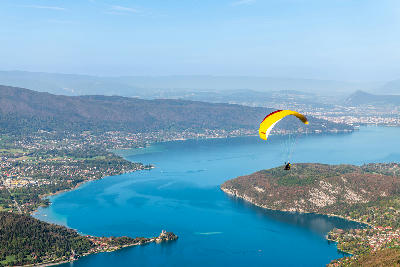 Paraglider flying above Lake Annecy with turquoise water, forested hills, and lakeside towns in the French Alps