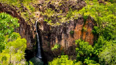 High-angle view of Tolmer Falls cascading down a narrow sandstone gorge into a dark plunge pool, surrounded by orange cliffs and lush green vegetation in Litchfield National Park, Northern Territory, Australia.