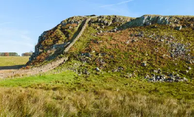 Hadrian's Wall ascending the rocky hillside at Peel Crags in Northumberland, England, surrounded by grassland, autumn bracken and exposed stone beneath a blue sky.