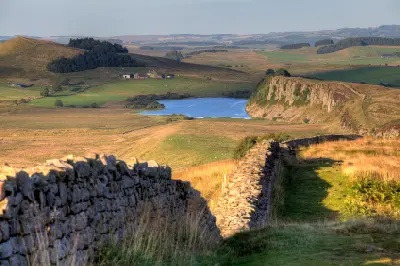 View east from Winshields along Hadrian's Wall towards Crag Lough, with rocky Highshield Crags and rolling Northumberland countryside in the distance.
