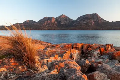 The Hazards mountain range glowing in warm evening light across calm water, with orange-lichen-covered granite rocks in the foreground, Freycinet National Park, Tasmania.