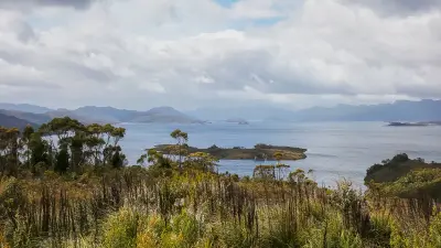Lake Pedder with native vegetation, small islands and layered mountain ranges beneath cloudy skies in southwest Tasmania, Australia.