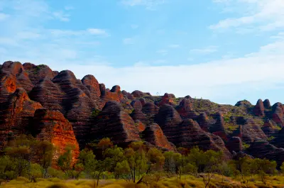 Distinctive orange-and-black striped sandstone domes of the Bungle Bungle Range rise above sparse savannah vegetation beneath a blue sky.