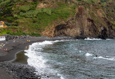 Swimmers and visitors on the black volcanic sand of Mosteiros Beach beneath coastal cliffs on São Miguel Island in the Azores.