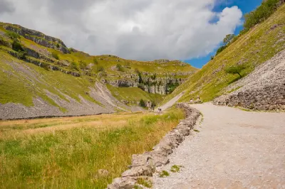 Limestone cliffs and grassy slopes surrounding a walking path near Malham Cove, with hikers exploring the Yorkshire Dales landscape.