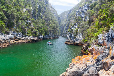 Storms River Gorge with steep rocky cliffs, emerald water and a small boat in Tsitsikamma National Park, South Africa.