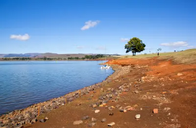 Rocky shoreline exposed by lower water levels at Midmar Dam, with calm water and rolling hills in the background.