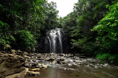 Wide landscape view of Ellinjaa Falls surrounded by dense rainforest, with visitors beside the waterfall and a rocky stream in the foreground.