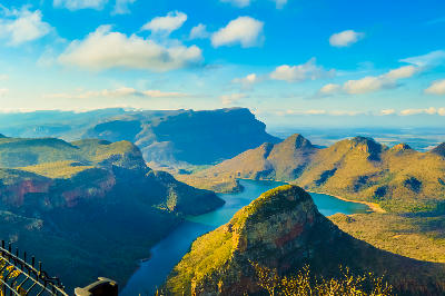 Panoramic view of Blyde River Canyon and the Three Rondavels in Mpumalanga, South Africa, with the winding Blyde River, towering cliffs, rounded mountain peaks and expansive canyon scenery beneath a bright blue sky.