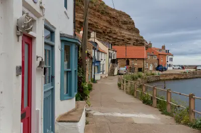 Colourful cottage doors and windows beside a waterfront path in Staithes, North Yorkshire, with traditional stone buildings and a towering coastal cliff.