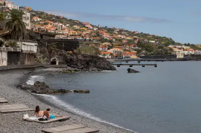 Young couple sitting on a pebble beach in Santa Cruz, Madeira, with coastal buildings and hillside houses overlooking the Atlantic Ocean.