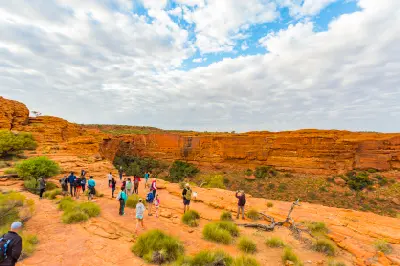 Tourists hiking across the rocky sandstone landscape at Kings Canyon in Central Australia, with dramatic red canyon cliffs, sparse vegetation and a partly cloudy blue sky.