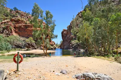 Unidentified people sit by the clear, calm waters of a scenic outback lake and waterhole, framed by sandy shores and tall eucalyptus trees under a clear blue sky.