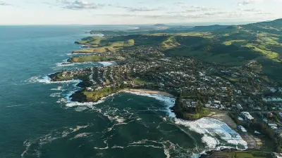 Sweeping aerial view across Kiama’s beaches, rocky headlands and coastal town toward rolling green hills and the distant Illawarra coastline.