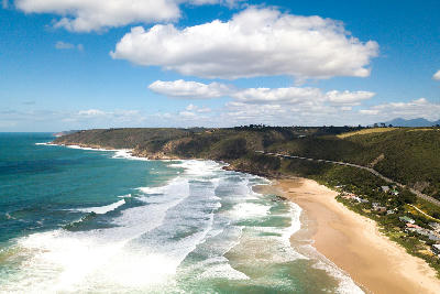 Aerial view of Wilderness Beach on South Africa’s Garden Route, showing rolling surf, a wide sandy shoreline and rugged coastal cliffs stretching into the distance.
