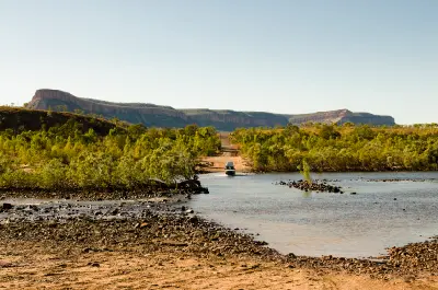 Four-wheel-drive crosses the shallow Pentecost River with the rugged red escarpments of the Cockburn Range stretching across the background.
