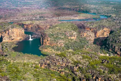 Aerial view of Mitchell Falls cascading into deep pools surrounded by rugged sandstone gorges, with the Mitchell River winding across the plateau beyond.