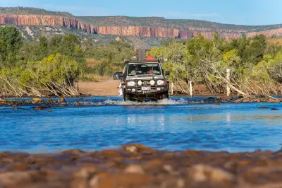Four-wheel-drive vehicle splashes through the shallow Pentecost River with the red cliffs of the Cockburn Range in the background.