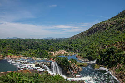 Water flowing over the Hartbeespoort Dam spillway into the Crocodile River, surrounded by green valleys and rocky hills in the Magaliesberg region of South Africa.