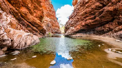 A calm waterhole reflecting the blue sky as it cuts through dramatic, layered red-orange rock cliffs at Simpsons Gap or Rungutjirpa.