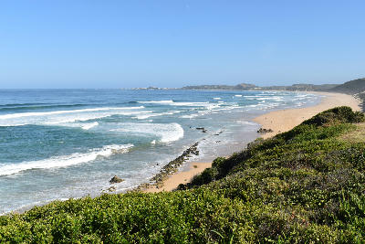 Panoramic view of Brenton Beach showing waves breaking along a long sandy coastline with coastal fynbos-covered cliffs near Knysna, South Africa.