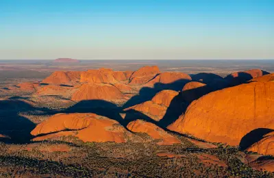 Aerial view of the red sandstone domes of Kata Tjuta in evening light, with Uluru visible on the distant horizon.
