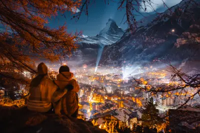 Couple overlooking illuminated Zermatt village at night with the Matterhorn rising above the valley in winter.