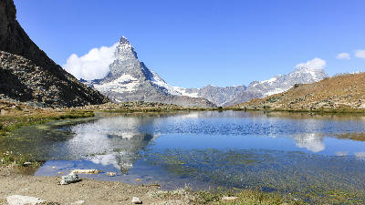 Matterhorn reflected in Riffelsee with shallow shoreline and alpine terrain under clear blue sky, Zermatt, Switzerland.