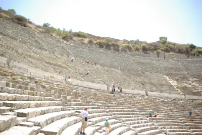 Tourists walking among the vast semicircular tiers of weathered stone seats inside the Great Theatre of Ephesus.