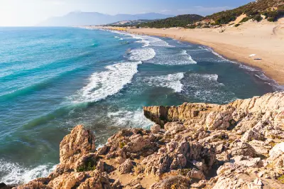 Elevated coastal view of Patara Beach, with turquoise waves rolling toward a long sandy shoreline, rugged limestone rocks in the foreground and hazy mountains in the distance.