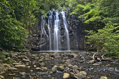 Frontal view of Ellinjaa Falls cascading over dark rocks into a rainforest pool, with a shallow rocky creek in the foreground near Millaa Millaa, Queensland.