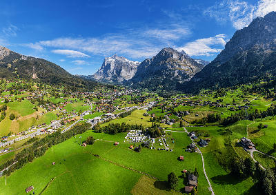 Aerial view of Grindelwald village with green meadows, winding roads, and dramatic Swiss Alps mountains under clear sky.
