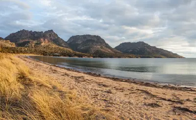 The Hazards mountain range viewed across Coles Bay, with a sandy shoreline and golden coastal grasses in the foreground, Freycinet National Park, Tasmania.