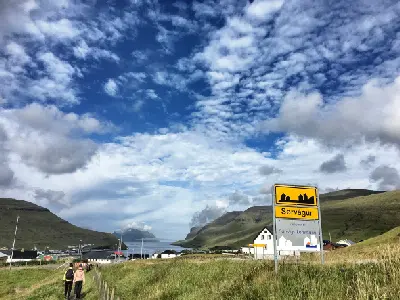 Roadside entrance sign for Sørvágur on Vágar Island, Faroe Islands, with two people walking toward the settlement and Mykines island visible across the fjord.