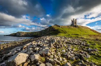 Dunstanburgh Castle ruins standing on a grassy coastal headland above rocky shores and sea cliffs in Northumberland, England, beneath dramatic storm clouds.