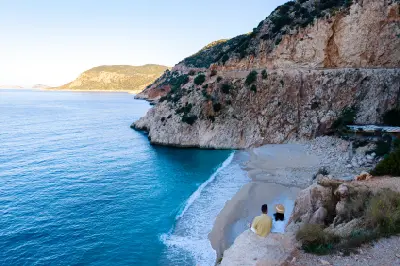 Two people sit together on a rocky overlook above Kaputaş Beach, watching turquoise waves reach the small sandy cove beneath steep limestone cliffs.