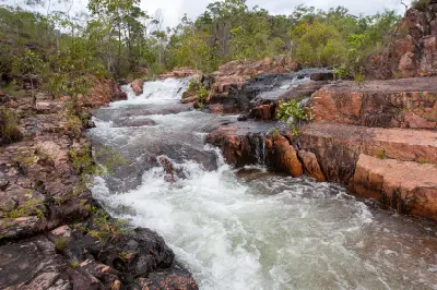 Fast-flowing stream cascading over reddish sandstone rocks and shallow ledges above Tolmer Falls, surrounded by green woodland in Litchfield National Park, Northern Territory, Australia.