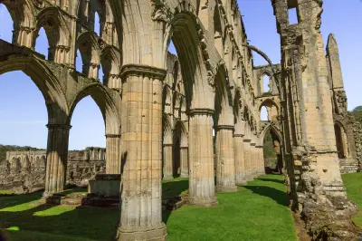 Interior view of Rievaulx Abbey ruins showing massive medieval stone columns, pointed Gothic arches and sunlit green lawns in North Yorkshire.