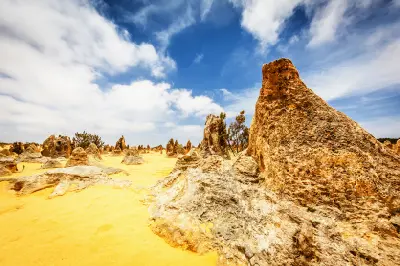 Large weathered limestone pinnacles rise from yellow sand beneath a blue and cloud-filled sky.