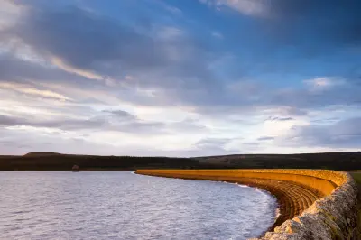 Curved stone embankment beside Kielder Water reservoir in Northumberland, with rippling water, distant forest and a blue-grey evening sky.