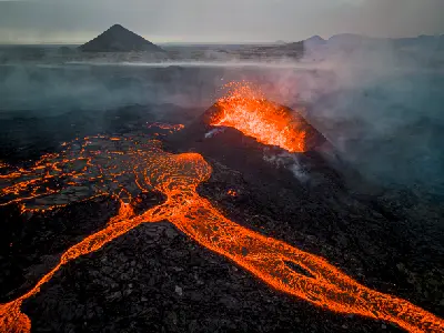 Drone view of an active volcanic crater with bright orange lava flowing across the dark landscape on the Reykjanes Peninsula, Iceland.