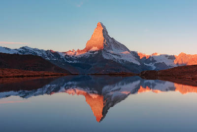 Matterhorn peak illuminated by sunrise and reflected in Stellisee lake near Zermatt, Switzerland, with calm water and clear sky.