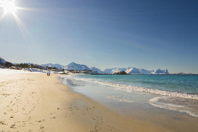 Bøvær Beach Senja Norway with long sandy shoreline, clear turquoise water, coastal houses and snow covered mountains under bright sunlight.