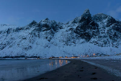 Dark sand shoreline of Ersfjord Beach with reflections and illuminated houses beneath steep snowy mountains