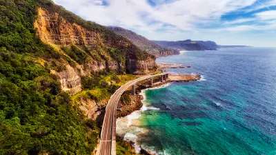 Aerial view of Sea Cliff Bridge sweeping beneath steep sandstone cliffs beside turquoise Pacific waters on the Illawarra coast.